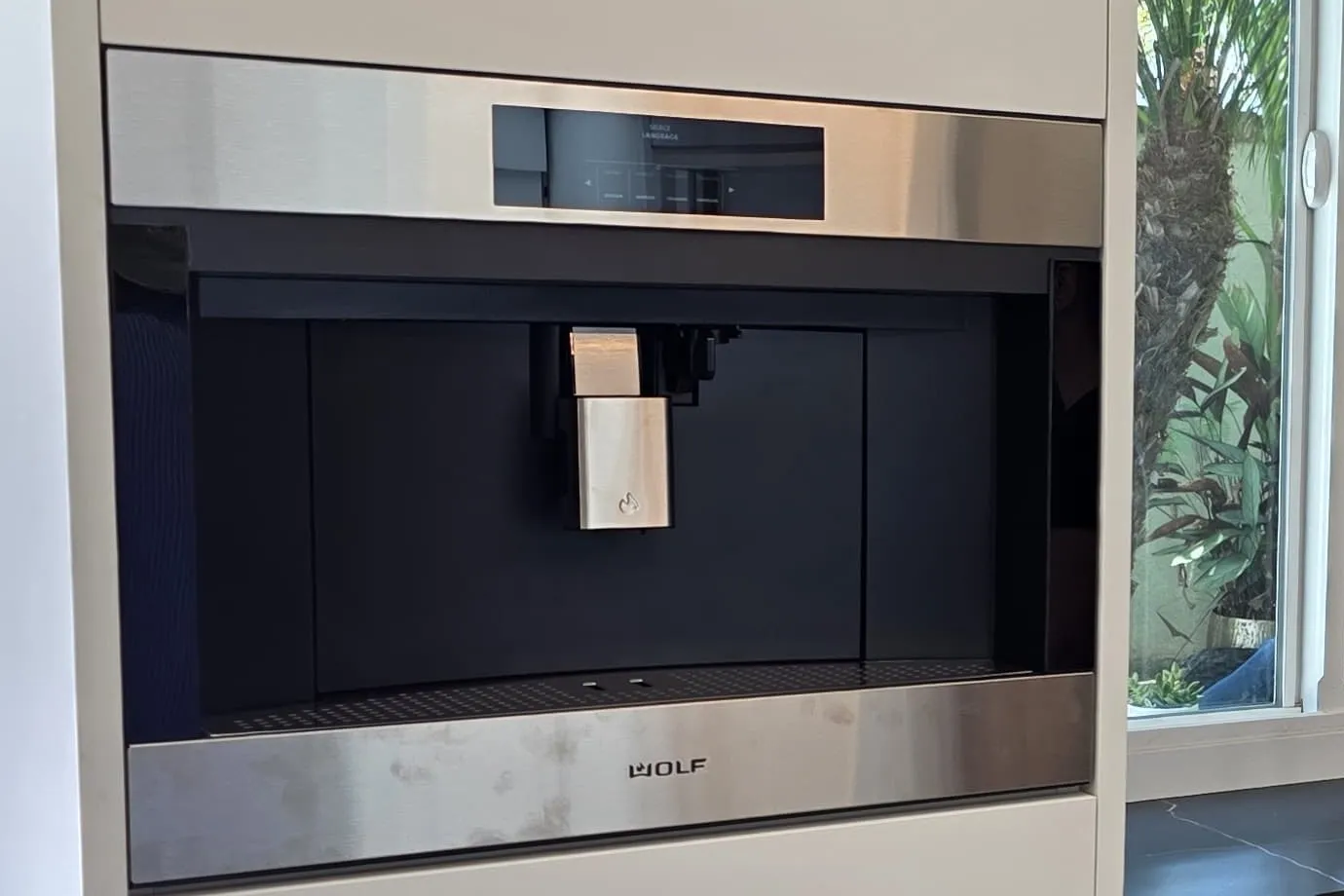 Mid-install kitchen detail: a built-in Wolf coffee system set into a handleless white slab cabinet column, beside a black marble-look counter and a garden window